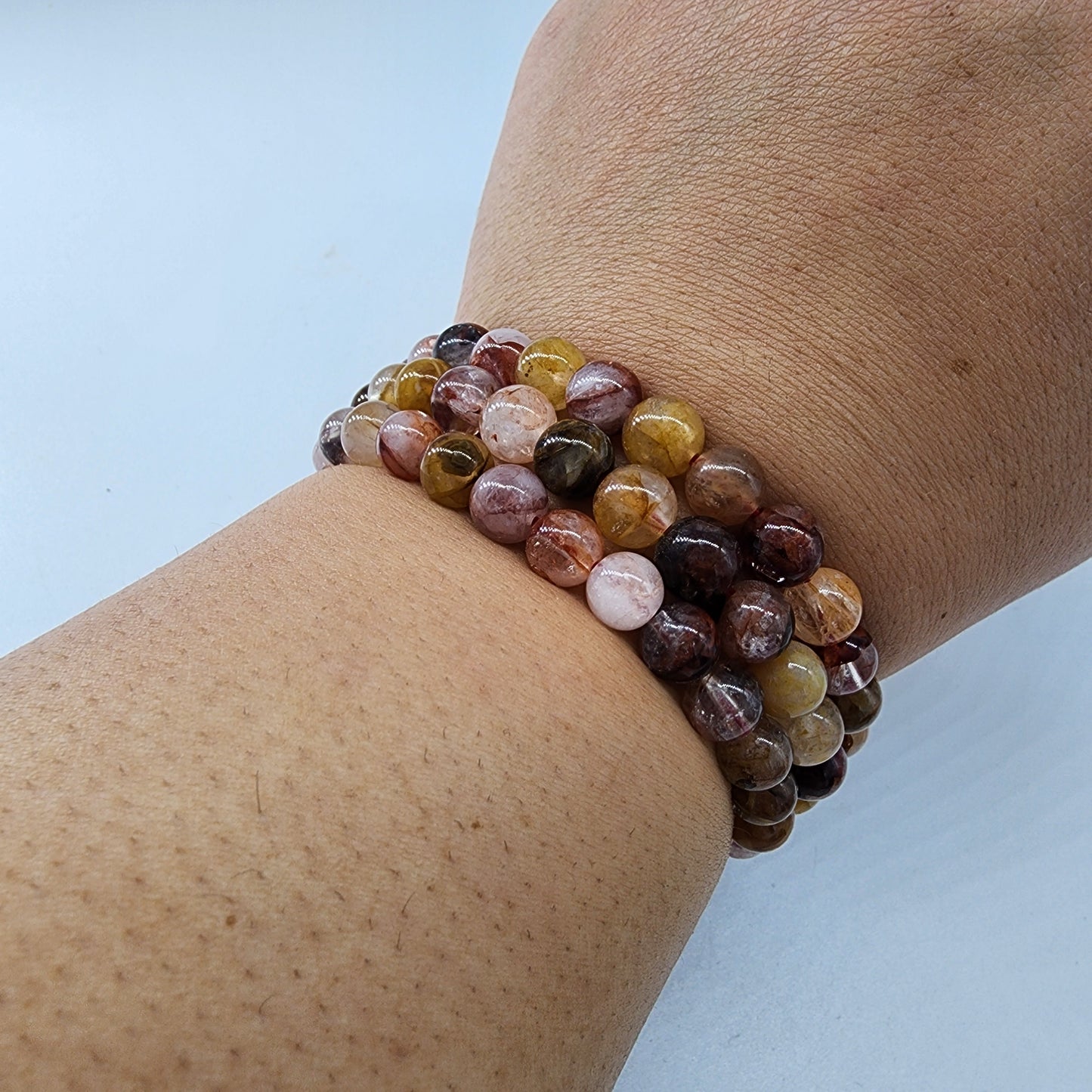 A person's wrist adorned with three Hematoid Quartz Crystal Bracelets from The Crystalary, featuring polished, multicolored round stones in shades of brown, yellow, pink, and black. These elegant adornments symbolize spiritual growth and higher awareness against a plain white background.