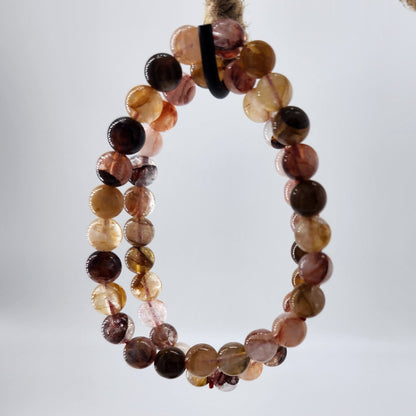 A close-up of The Crystalary's Hematoid Quartz Crystal Bracelet, featuring round, polished stones in various shades of brown, orange, and pink. The bracelet is elegantly draped on a twine-wrapped circle, set against a blurred white background to symbolize spiritual growth and higher awareness.