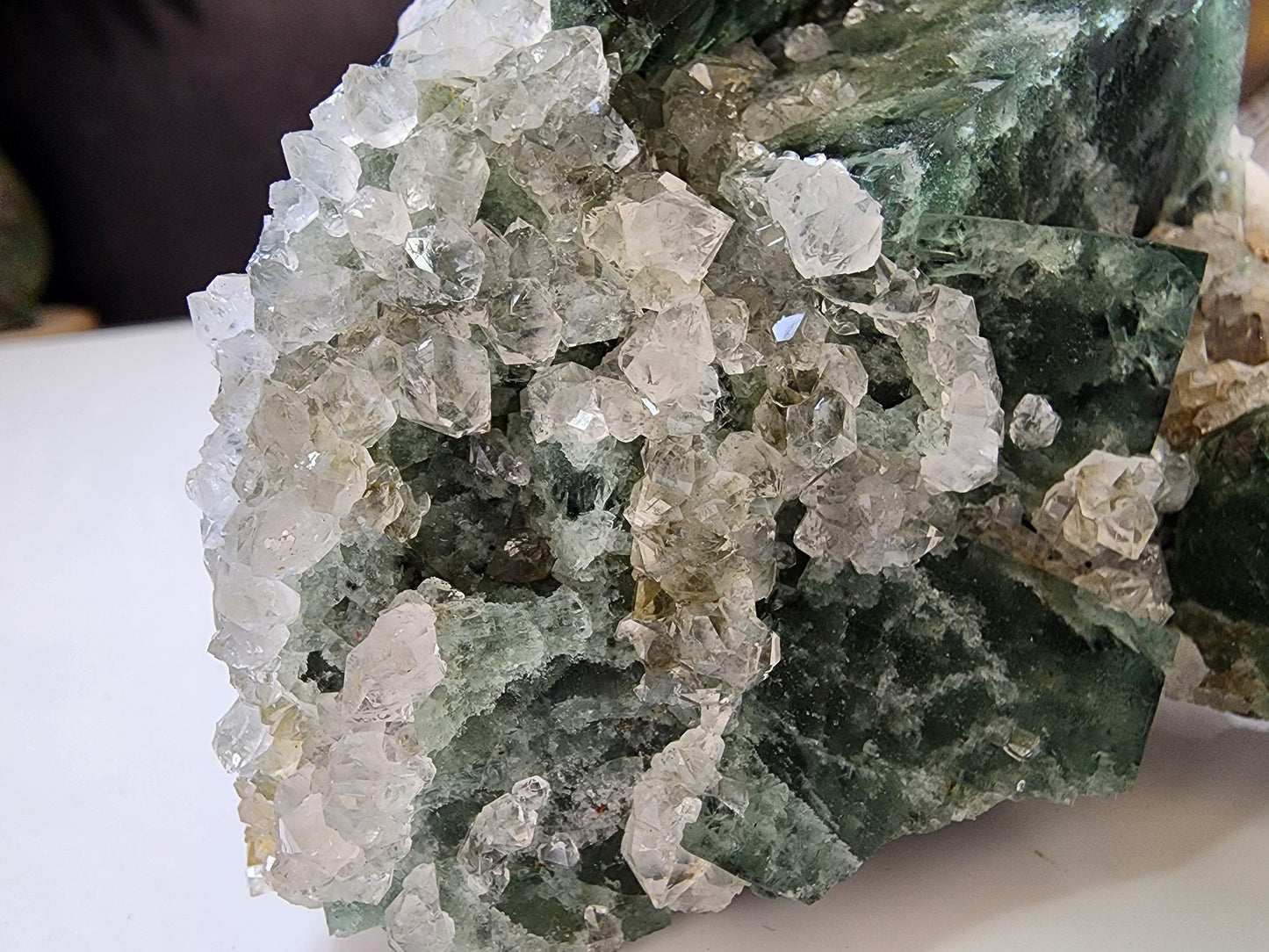 A close-up image of the Hidden Forest Pocket Fluorite with Quartz from The Crystalary, sourced from the Diana Maria Mine in Weardale, showcases a rough green mineral cluster. The green mineral is predominantly covered with clear, densely packed hexagonal fluorite crystals intergrown with quartz, giving the formation a jagged and sparkling appearance.