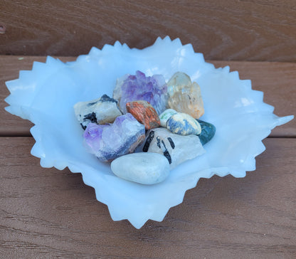 An Indiana Glass Diamond Point Ruffled Candy/Trinket Dish from The Crystalary holds various polished stones and crystals, including amethyst, quartz, and other multicolored minerals. This crystal display is beautifully arranged on a wooden surface.