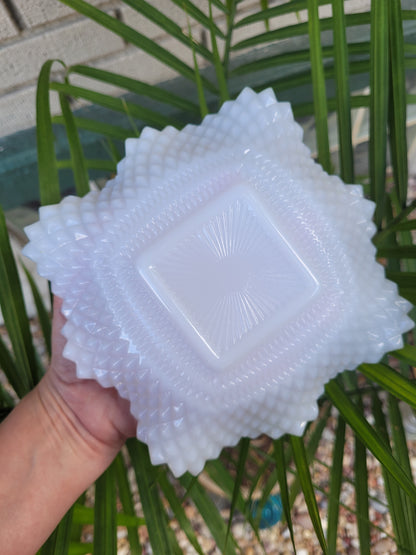 A hand holding The Crystalary's Indiana Glass Diamond Point Ruffled Candy/Trinket Dish, featuring its white, intricately designed surface and textured edges, is set against a backdrop of green palm leaves and a light-colored wall.