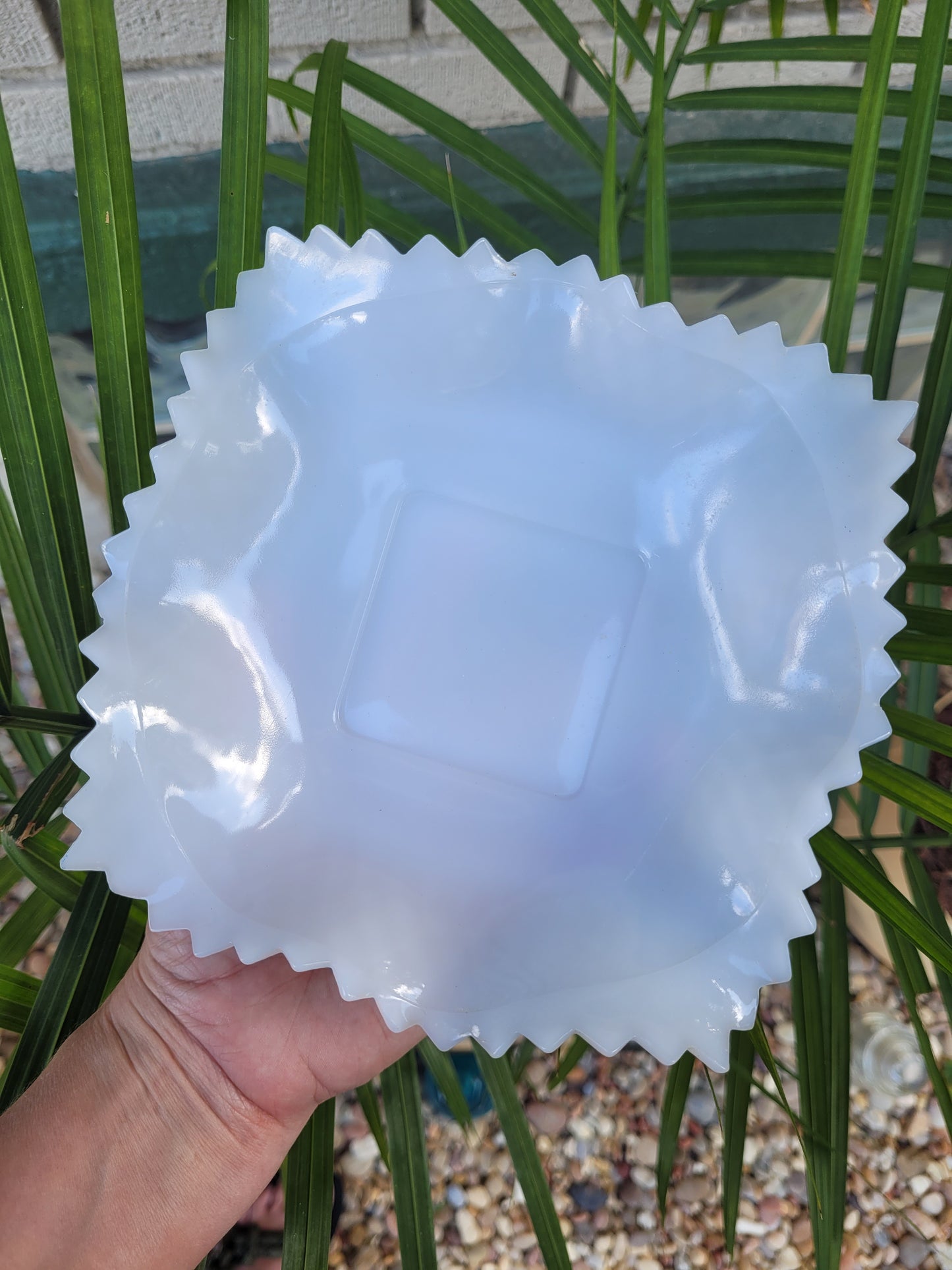 A hand is holding an Indiana Glass Diamond Point Ruffled Candy/Trinket Dish from The Crystalary, featuring a flexible, translucent, blue silicone cover with wavy, serrated edges. The background showcases green palm fronds and gravel, creating an outdoor ambiance that complements the aesthetic of vintage dishes.