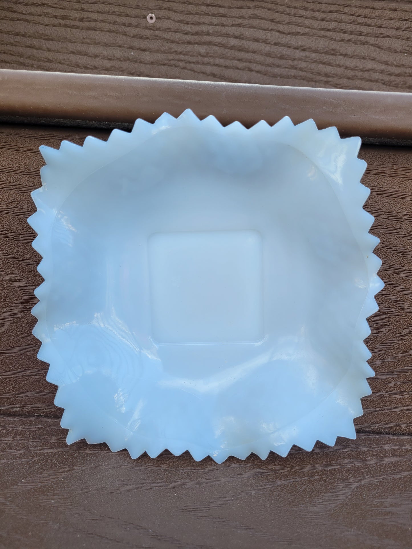 A square, translucent white glass dish from The Crystalary, known as the Indiana Glass Diamond Point Ruffled Candy/Trinket Dish, features wavy, ruffled edges and a glossy finish. Reminiscent of vintage designs, it boasts a smooth, flat center and a decorative scalloped border against a wooden background.