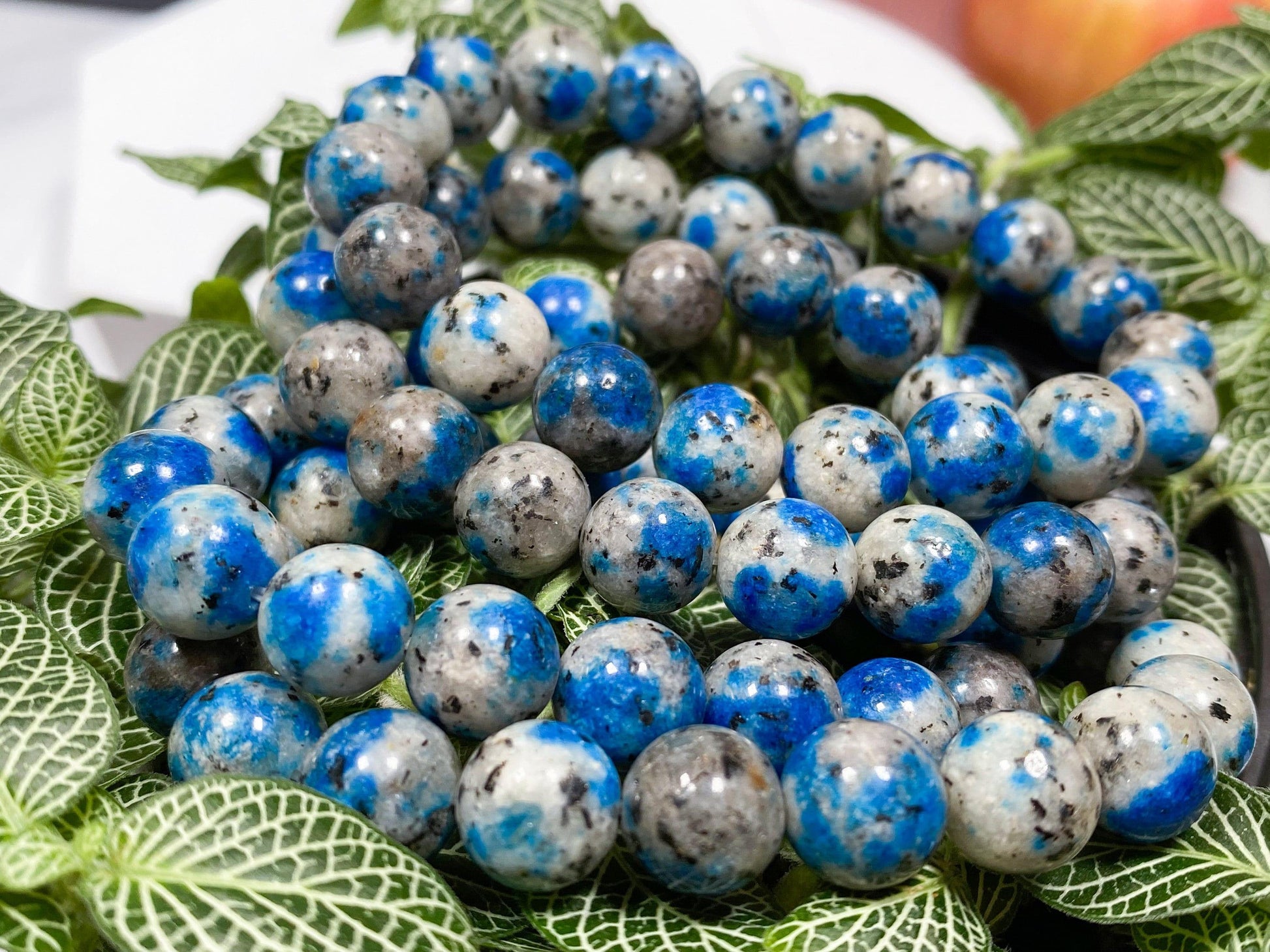 Image of a K2 Crystal Bracelet made of polished blue and gray speckled K2 Jasper stones, resting on foliage with green and white leaves. In the background, an apple is partially visible, highlighting the natural elegance of gemstone bracelets by The Crystalary.