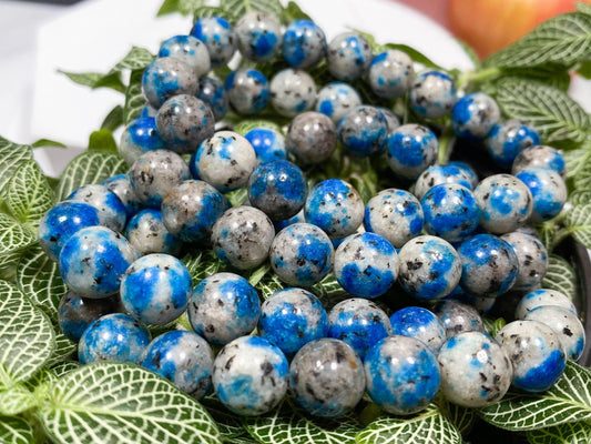 Image of a K2 Crystal Bracelet made of polished blue and gray speckled K2 Jasper stones, resting on foliage with green and white leaves. In the background, an apple is partially visible, highlighting the natural elegance of gemstone bracelets by The Crystalary.