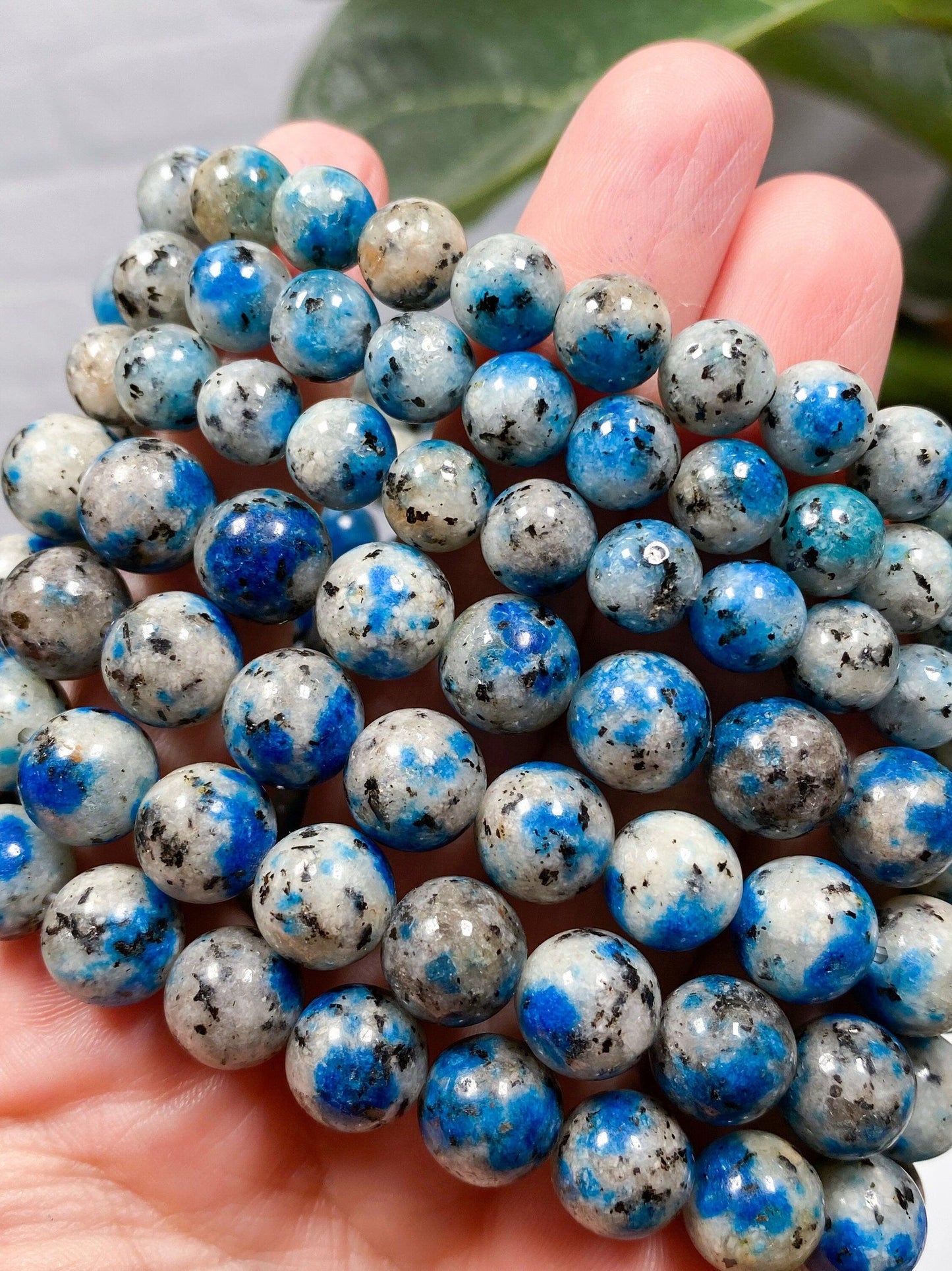 A person's hand holds a cluster of smooth, round beads, marbled with shades of blue, gray, white, and black. These K2 Crystal Bracelets from The Crystalary glimmer softly against the blurred green leaf in the background.