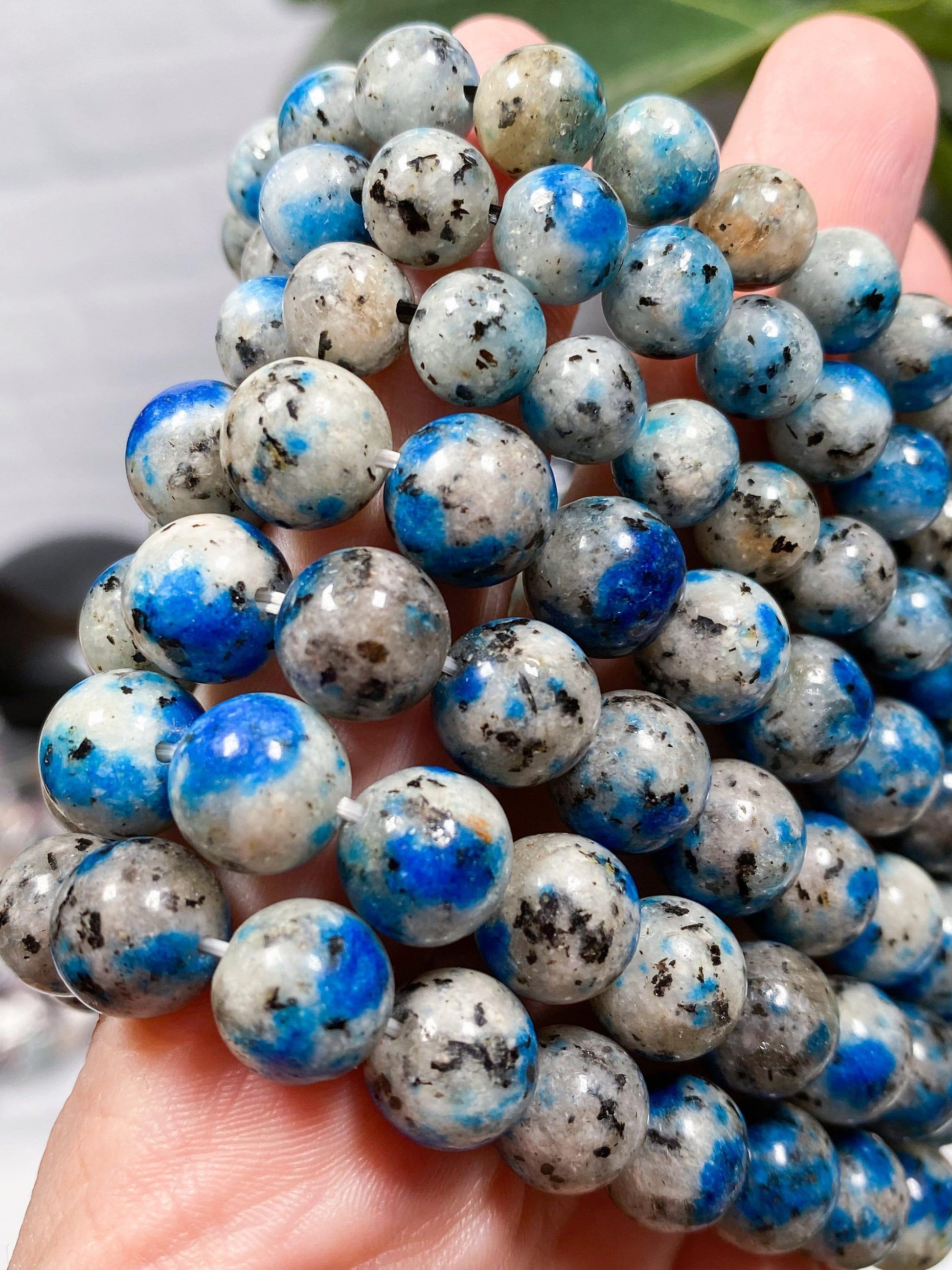 Close-up image of a hand holding multiple strands of round, polished K2 Crystal beads from The Crystalary's K2 Crystal Bracelet collection. The beads are predominantly light gray with vibrant blue and some darker speckles, giving them a striking marbled appearance. Perfect for creating unique gemstone bracelets, the background is slightly blurred.