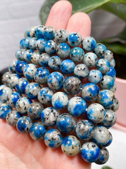 A hand holding a string of The Crystalary’s K2 Crystal Bracelet. The polished beads feature a marbled pattern with shades of blue, gray, and black. A blurred plant and part of a pink pot are visible in the background.