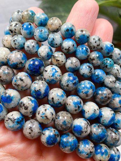 A close-up of a hand holding a bunch of polished round beads from The Crystalary's K2 Crystal Bracelet. The beads are predominantly grey with striking blue azurite patches and black speckles, creating a visually appealing contrast often seen in exquisite gemstone bracelets. The background is slightly blurred, putting focus on the beads from the K2 Crystal Bracelet.