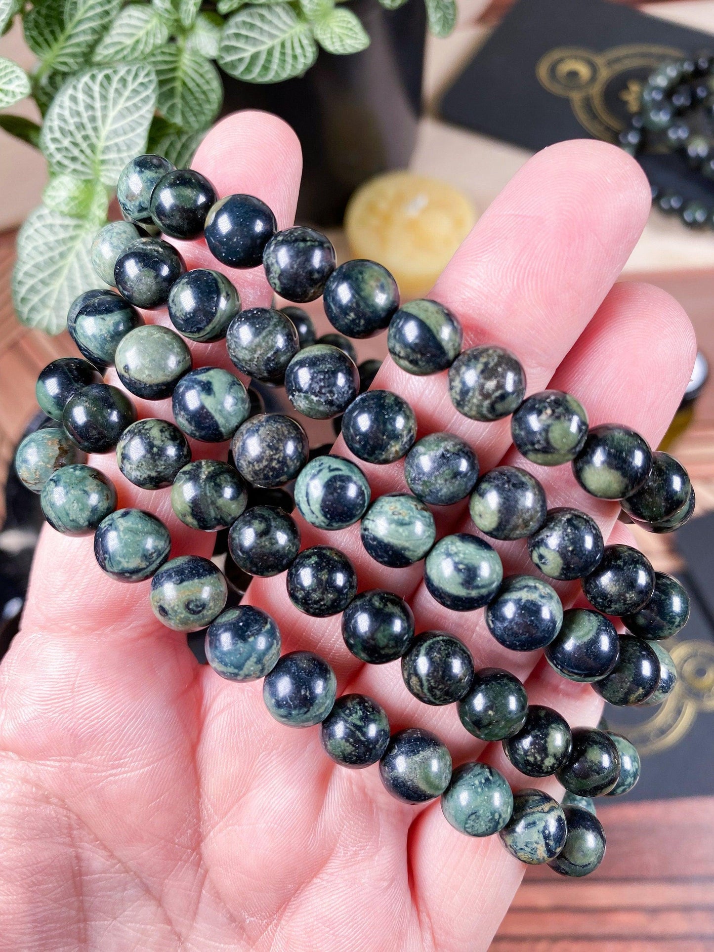 Close-up of a hand holding a strand of polished, round Kambaba Jasper (Green Stromatolite) beads from The Crystalary's 7.5-inch crystal bracelet, featuring a mix of dark green and black hues. The background shows a few plants and additional strands of beaded bracelets.