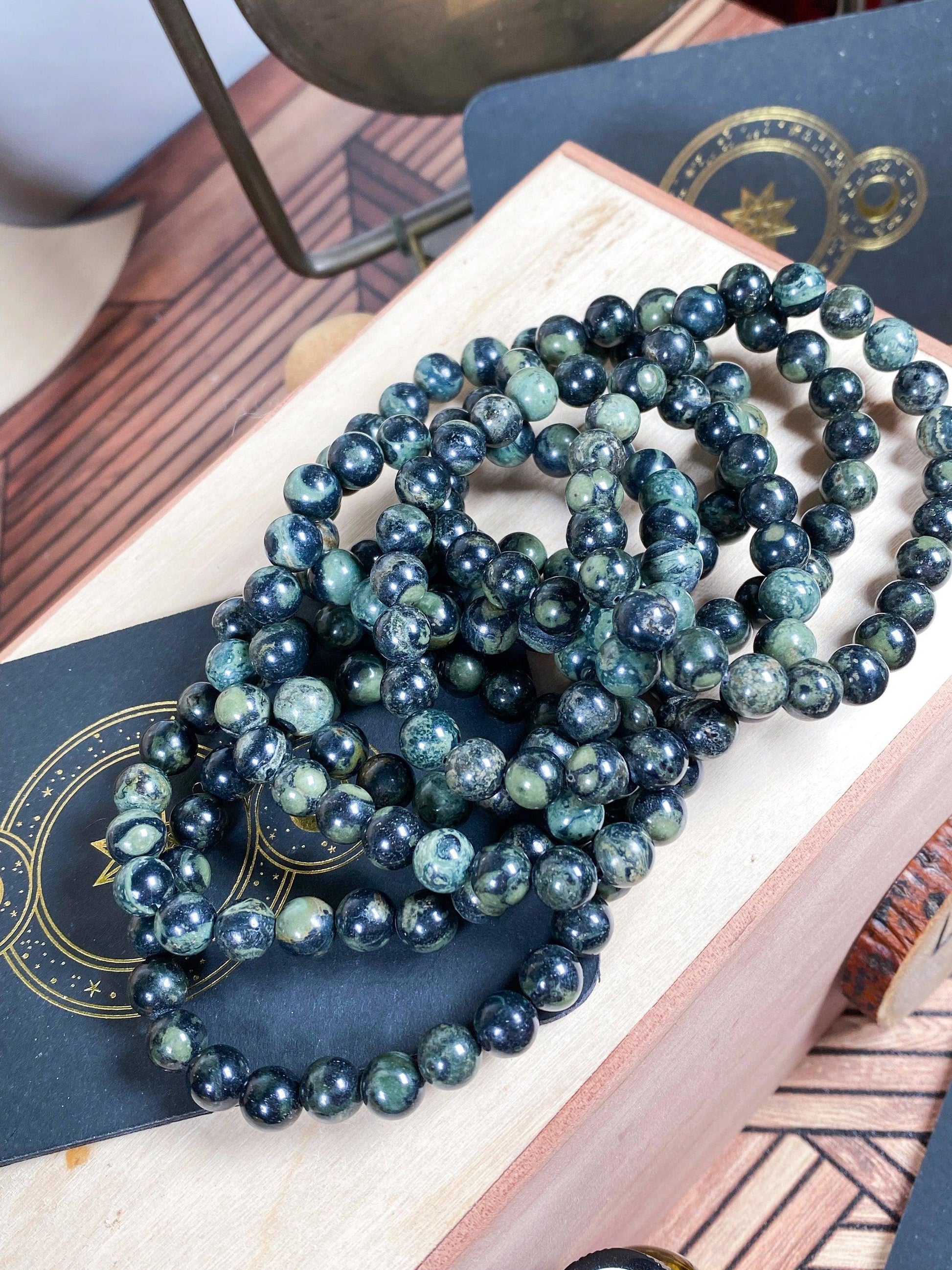 A collection of 7.5-inch Kambaba Jasper (Green Stromatolite) Crystal Bracelets by The Crystalary is displayed on a wooden box. Each bracelet features round, polished beads with slight variations in shade and texture, ranging from dark green to black. In the background, gold and black star-patterned cards are partially visible.