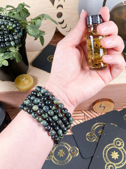 Wearing several of The Crystalary's Kambaba Jasper (Green Stromatolite) Crystal Bracelets, a hand gently holds a small vial filled with amber-colored liquid. In the background, tarot cards, a small candle, a plant, and a bowl containing more beaded bracelets set the scene.