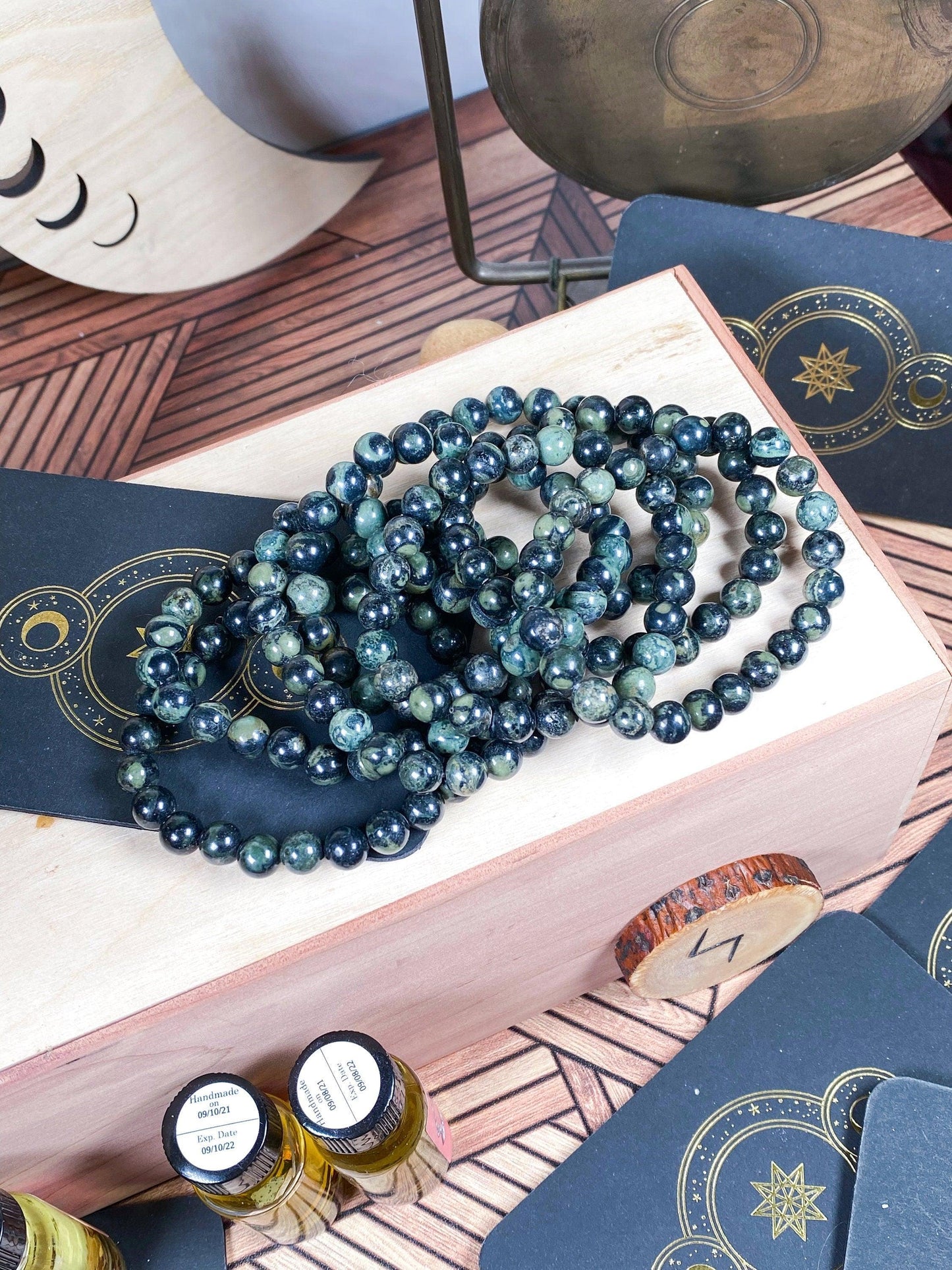 A cluster of dark beaded bracelets from The Crystalary, including the Kambaba Jasper (Green Stromatolite) Crystal Bracelet- 7.5in, lay on a wooden box adorned with a numeric "77" tag. Surrounding the box are black cards featuring gold geometric star designs and small bottles. The background is a patterned surface with additional decorative items.