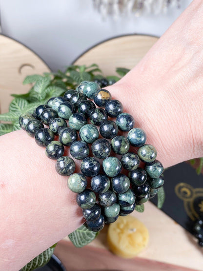 A hand adorned with five stacked crystal bracelets, including The Crystalary's 7.5-inch Kambaba Jasper (Green Stromatolite) Crystal Bracelet made of polished green and black gemstone beads. The background showcases some greenery and blurred objects, creating an earthy, natural vibe.