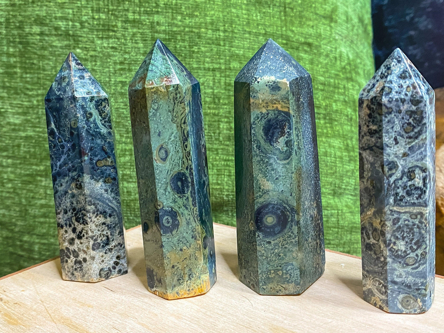 Four polished Kambaba Jasper (Green Stromatolite) Crystal Points from The Crystalary stand upright on a light-colored wooden surface, exhibiting varying patterns and shades of greenish-blue. The backdrop features a green fabric and a dark-colored area, contrasting beautifully with the crystals' intricate designs.