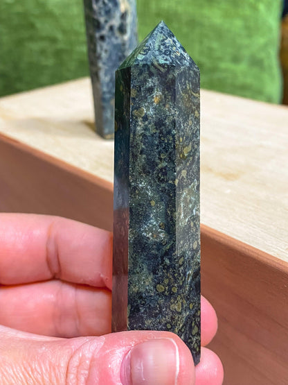 A person's hand holding a polished Kambaba Jasper (Green Stromatolite) Crystal Point from The Crystalary, showcasing intricate patterns and various shades. The background reveals a wooden surface with another healing crystal in the distance.