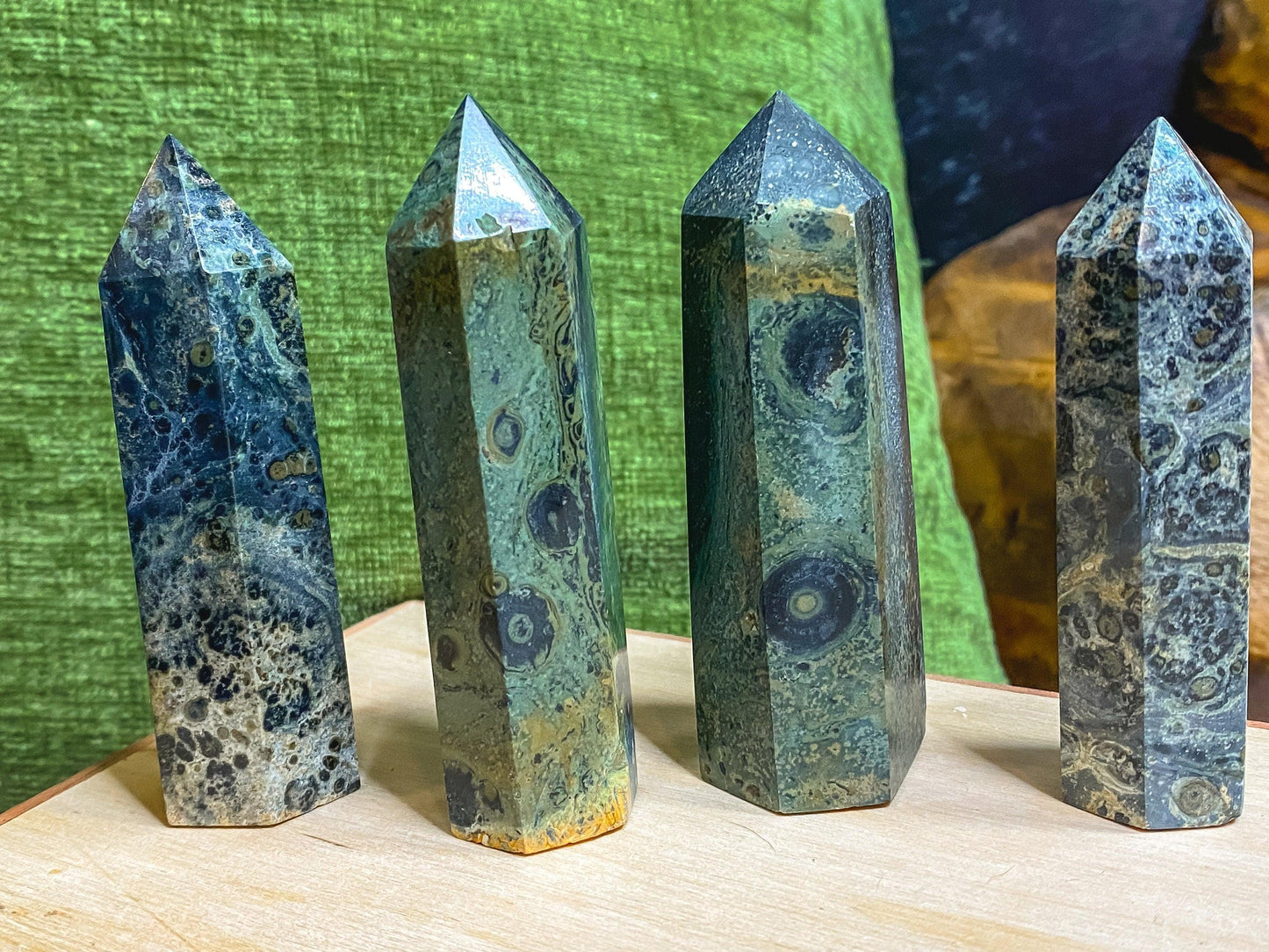 Four polished Kambaba Jasper (Green Stromatolite) Crystal Points from The Crystalary stand upright on a light-colored wooden surface. These healing crystals feature distinct green and black swirling patterns and are positioned against a green fabric backdrop with a darker background area.