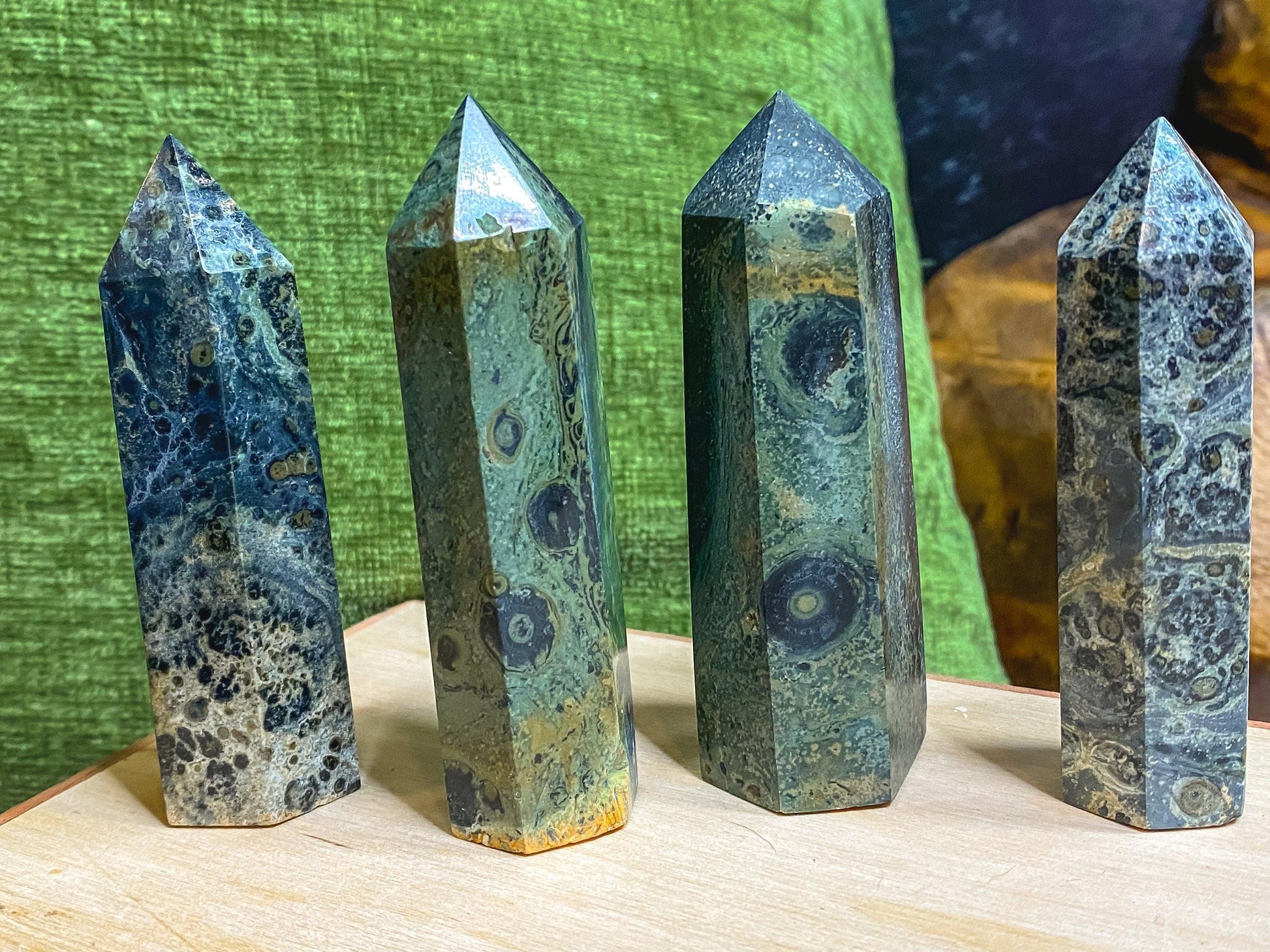 Four polished Kambaba Jasper (Green Stromatolite) Crystal Points from The Crystalary stand upright on a light-colored wooden surface. These healing crystals feature distinct green and black swirling patterns and are positioned against a green fabric backdrop with a darker background area.