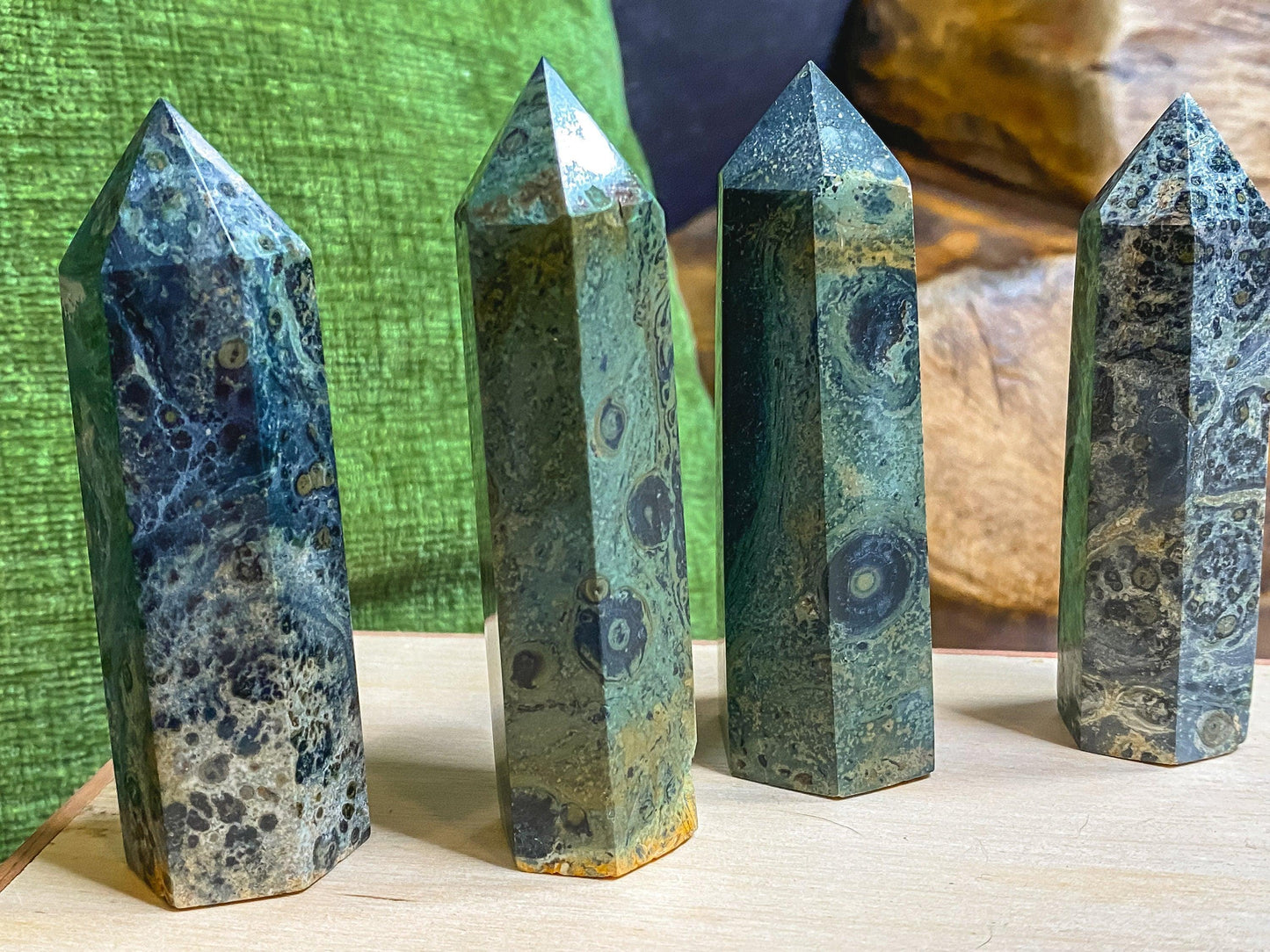 Four polished Kambaba Jasper (Green Stromatolite) Crystal Points from The Crystalary are arranged vertically on a wooden surface. In the background, a green cushion is situated on the left and a brown, textured object on the right. These healing crystals exhibit intricate patterns and visible variations in color.