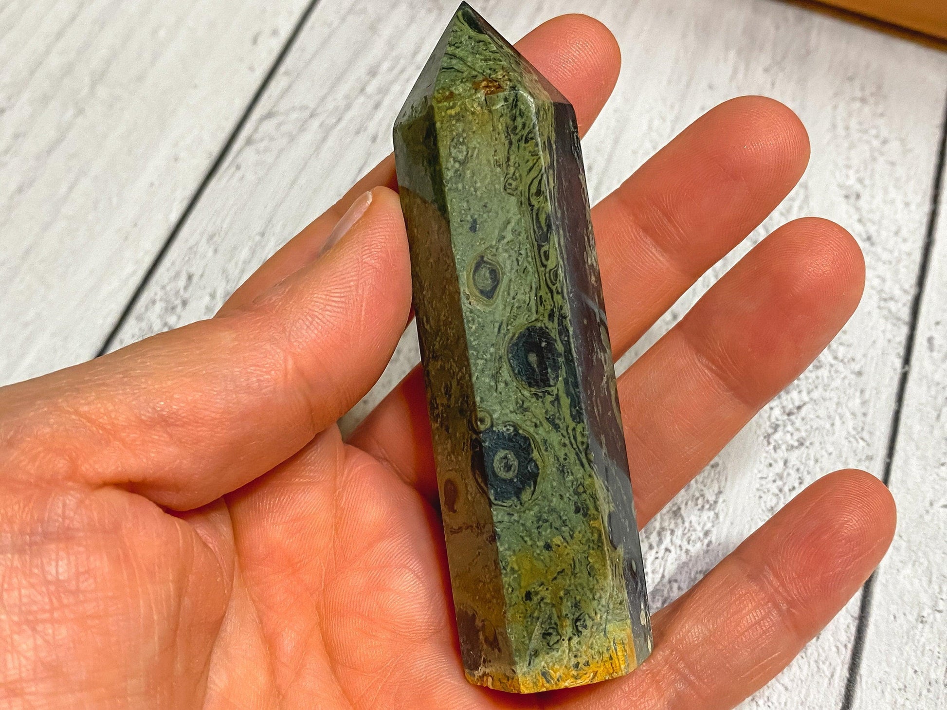 A hand holding a polished Kambaba Jasper (Green Stromatolite) Crystal Point from The Crystalary, featuring swirling patterns in shades of green, black, and yellow with circular markings visible on its surface. The crystal has a pointed tip, and the background is a wooden, whitewashed surface.