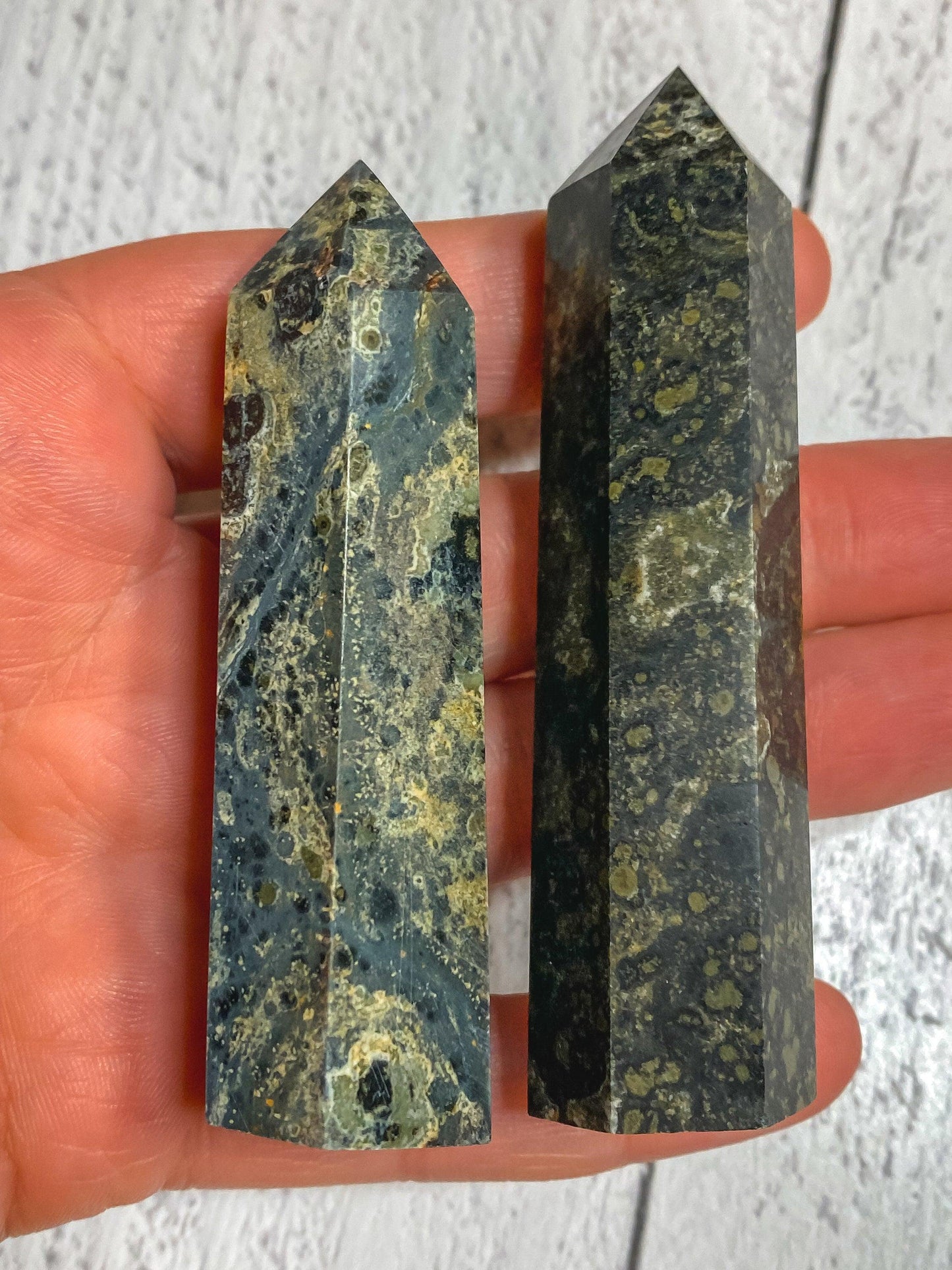 A hand holding two polished Kambaba Jasper (Green Stromatolite) Crystal Points by The Crystalary, displaying intricate patterns and varied shades of green and black. These healing crystals rest against a white, textured background.