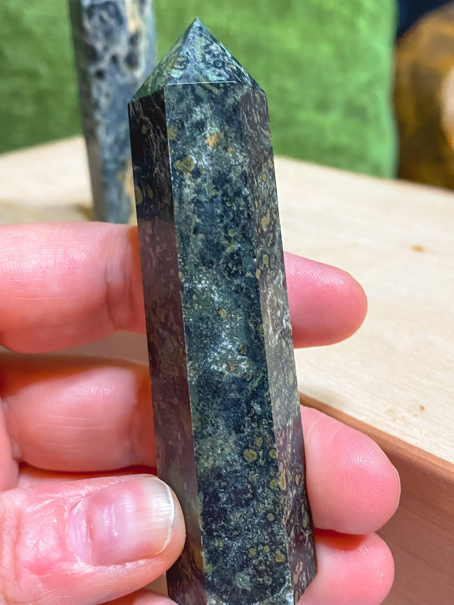 A hand holds a Kambaba Jasper (Green Stromatolite) Crystal Point from The Crystalary. The crystal point features a pointed tip and a speckled pattern, showcasing visible textures and color variations in shades of dark green and black. Another similar healing crystal is blurred in the background on a light wooden surface.