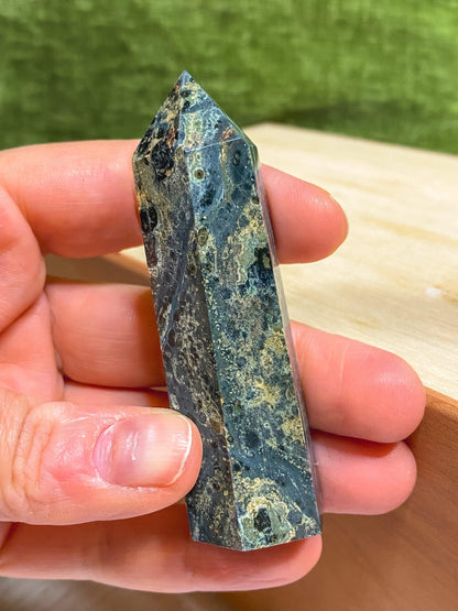 A hand holding a polished Kambaba Jasper (Green Stromatolite) Crystal Point from The Crystalary reveals intricate patterns of lighter and darker shades, showcasing its healing crystal properties. The background features a wooden surface with a green cloth or backdrop.