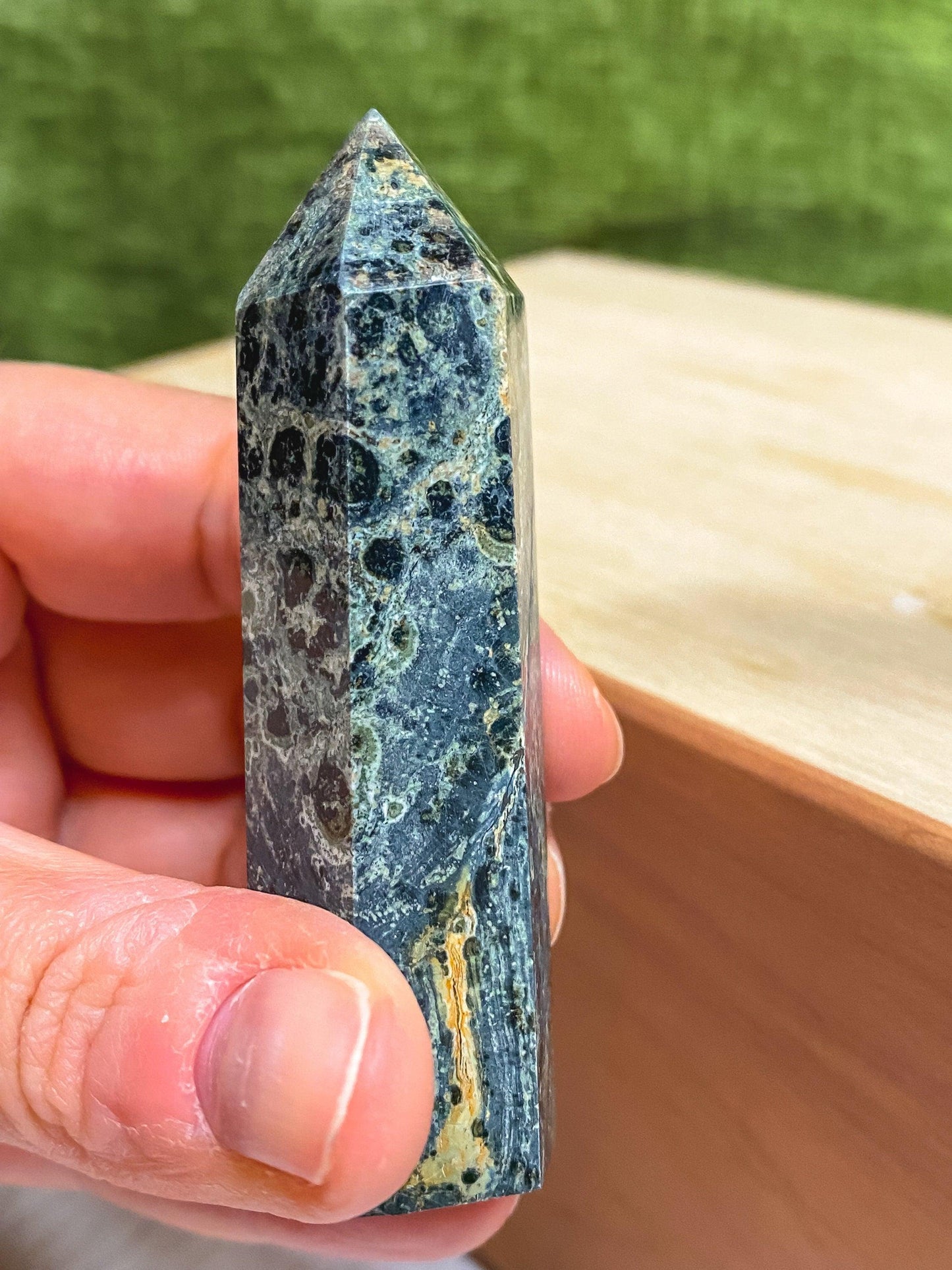 A hand holds an upright, hexagonal, polished crystal with a pointed tip. This Kambaba Jasper (Green Stromatolite) Crystal Point from The Crystalary has a greenish hue with intricate patterns of darker and lighter shades, resembling a semi-precious stone. The background is a blurred green and beige surface.