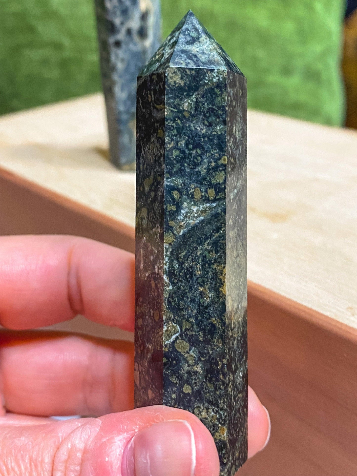 A hand holding an intricately patterned Kambaba Jasper (Green Stromatolite) Crystal Point by The Crystalary in front of a beige surface. The dark green stone has small, lighter spots and is smooth and glossy. Another similar healing crystal is visible in the blurred background.