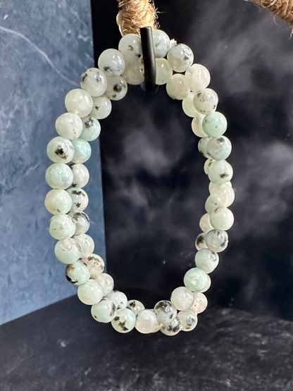 The Kiwi Jasper Crystal Bracelet from The Crystalary is elegantly displayed on a unique stand wrapped in twine. The stand curves upward from a black base, creating a striking contrast against the bracelet's pale green hue. As one of many gemstone bracelets designed to absorb negative energy, it complements the dark gray textured background perfectly.