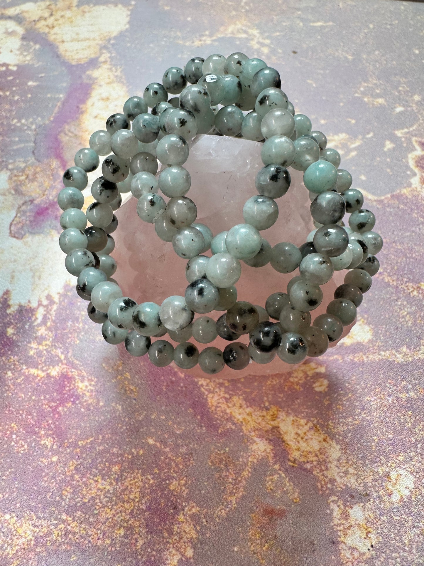 A Kiwi Jasper Crystal Bracelet by The Crystalary is displayed on a rough-cut, translucent cleansing crystal bracelet, set against a textured, multicolored pastel background. The polished, spherical green and black-speckled gemstones create an elegant and earthy accessory that may absorb negative energy.