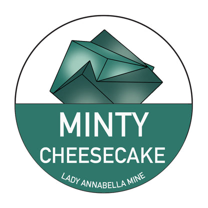 A logo featuring a green geometric crystal with a split background of green and white, capturing the vibrant hues of fluorite. Below the crystal, bold white text reads "MINTY CHEESECAKE." Smaller text at the bottom reads "LADY ANNABELLA MINE." Ideal for full-color stickers, these are part of The Crystalary's Lady Annabella Passport Stickers collection.