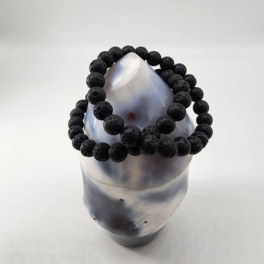 The Lava Rock Crystal Bracelet from The Crystalary features a series of round black beads elegantly encircling a smooth, conical gemstone. Renowned for its detoxification properties, the gemstone showcases a subtle gradient of white and bluish-gray hues, all set against a plain white background.