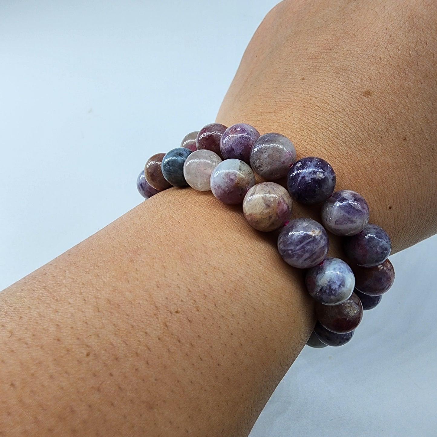 A wrist adorned with The Crystalary's Lavender Rose Quartz Crystal Bracelet showcases polished, round purple and Lavender Rose Quartz stones, celebrated as the stone of unconditional love, resting gracefully on a light background.