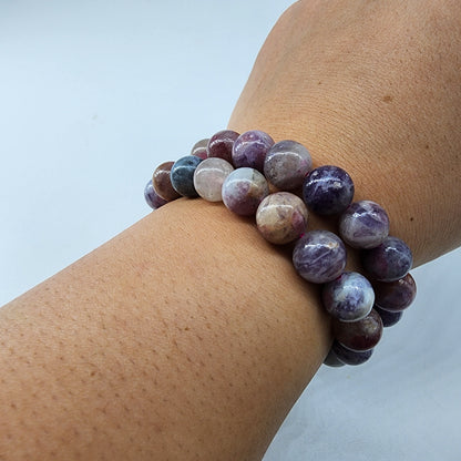 A wrist adorned with The Crystalary's Lavender Rose Quartz Crystal Bracelet showcases polished, round purple and Lavender Rose Quartz stones, celebrated as the stone of unconditional love, resting gracefully on a light background.