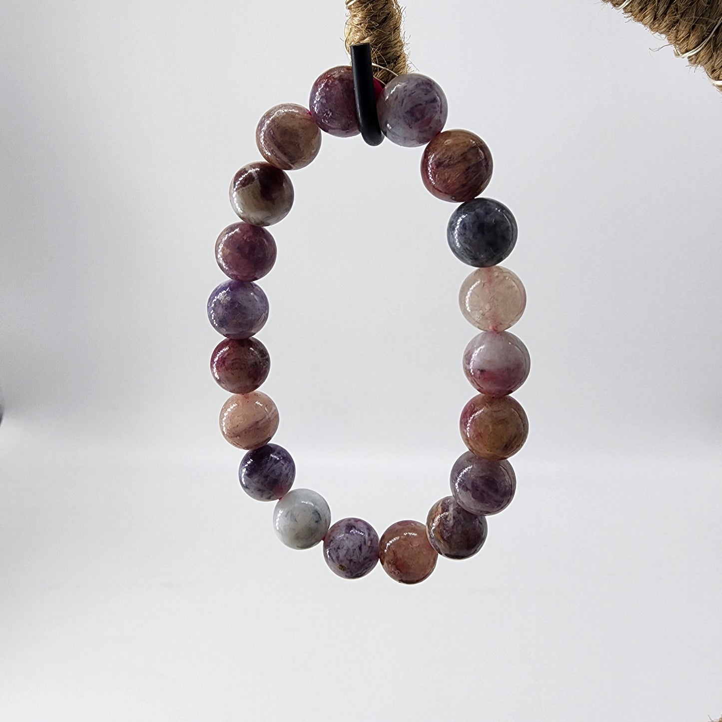 Effortlessly suspended in the air against a plain white backdrop is The Crystalary's Lavender Rose Quartz Crystal Bracelet, artfully crafted with Lavender Rose Quartz—a gemstone symbolizing unconditional love—and an array of round, multicolored gemstones in shades of purple, pink, and gray.