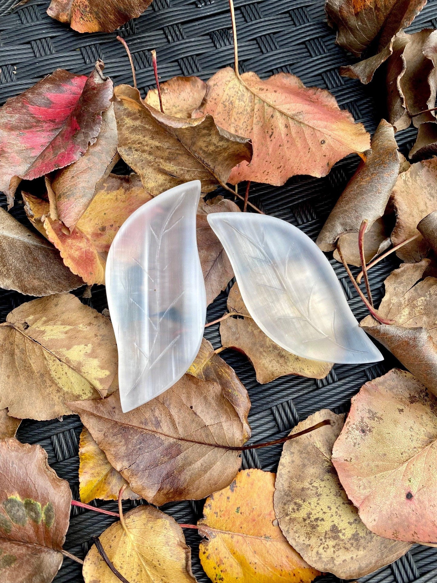 Two translucent, leaf-shaped soap bars lie amidst a scattering of autumn leaves on a textured, dark woven surface. The combination of natural and crafted elements creates an appealing contrast. A Leaf Selenite Charging Plate from The Crystalary complements the russet, gold, and brown tones that epitomize the fall season.