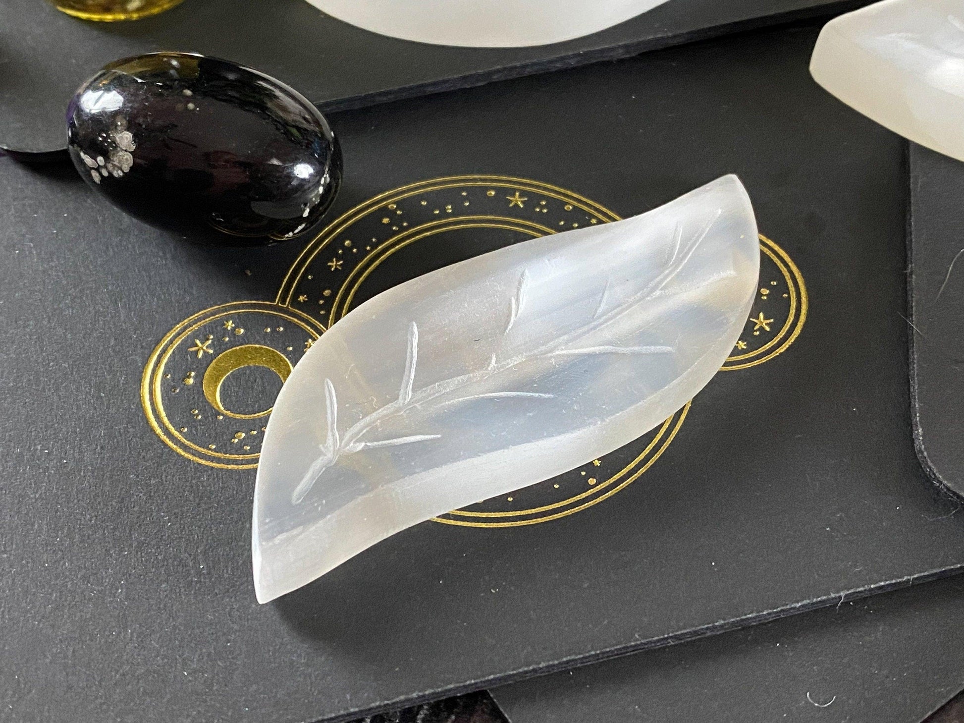 A Leaf Selenite Charging Plate from The Crystalary, featuring a translucent crystal carved in the shape of a leaf with intricately engraved stem and veins, is placed on a black surface adorned with a golden geometric design. To the left, there is also a polished black stone speckled with white, adding to the elegant natural charging display.