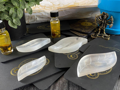 A table setup displays carved leaf-shaped crystal bowls, small bottles of golden oil, a small statue of a multi-armed deity, and a plant on The Crystalary's Leaf Selenite Charging Plate. Black tarot cards with gold designs are laid out beneath the items. A blue pot and stone wall are visible in the background.