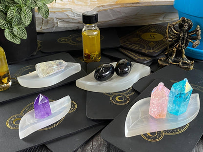 A selection of vibrant purple, pink, blue, and clear crystals displayed on The Crystalary's Leaf Selenite Charging Plate. These are accompanied by black and gold cards underneath each crystal. In the background, there is a small bottle of yellow oil, a mini statue, and verdant green leaves.