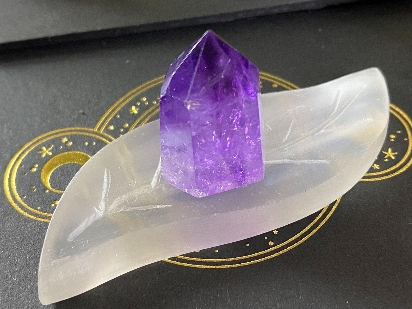 A polished purple crystal is resting on The Crystalary's Leaf Selenite Charging Plate, crafted from translucent selenite. The leaf-shaped dish, which resembles a natural charging surface, is placed on a dark background adorned with a golden celestial design featuring stars and a moon.