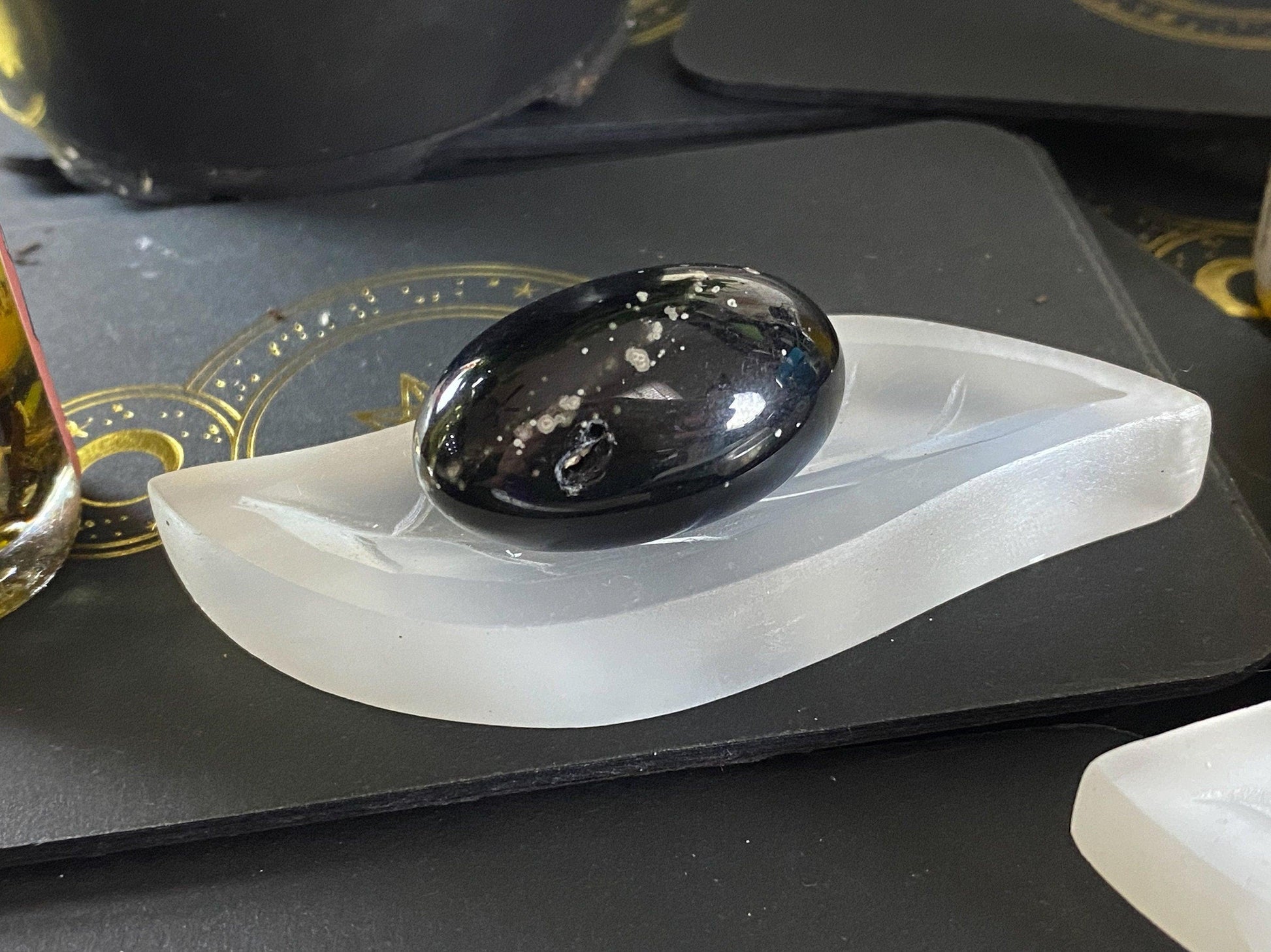 A polished black egg-shaped stone with white speckles sits on The Crystalary's Leaf Selenite Charging Plate, acting as a natural charging surface. The scene includes a black and gold printed surface underneath, partially visible.