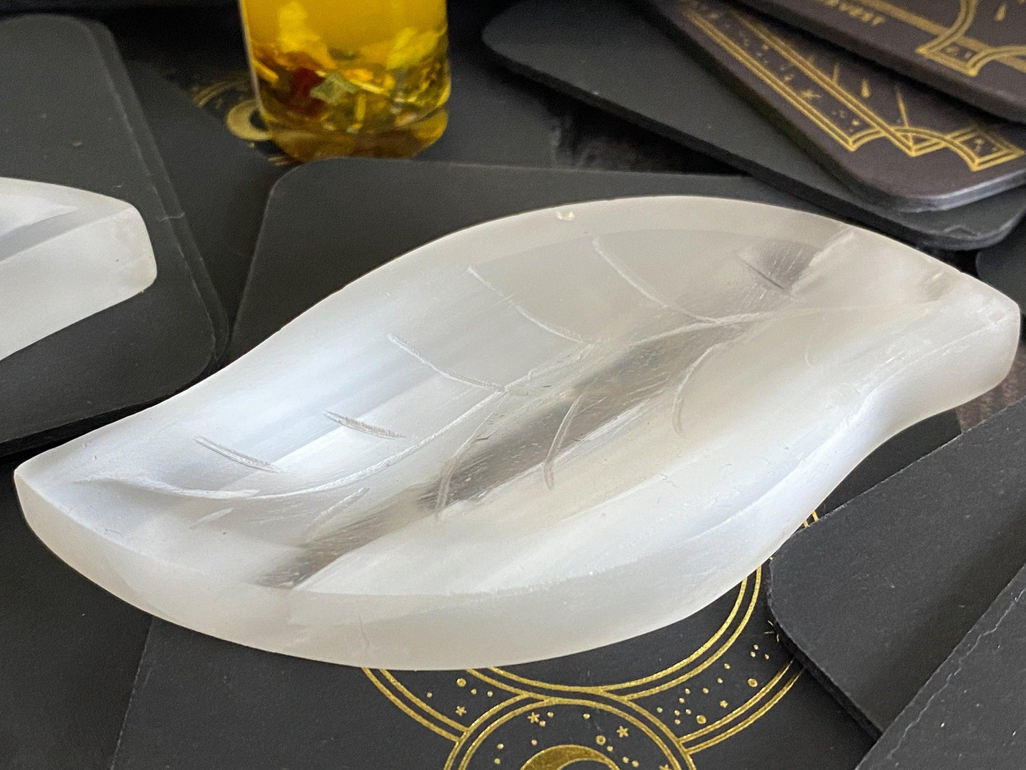 A smooth, translucent Leaf Selenite Charging Plate from The Crystalary rests on a dark surface adorned with intricate, golden patterns. Part of a yellow bottle with herbs inside is visible in the background. This beautifully crafted crystal serves as both art and a natural charging surface for your spiritual tools.