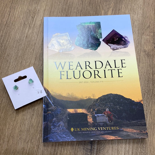 A book titled "LIVE- Ashley Pifer- 5/30/24" from The Crystalary, showcasing various fluorite mineral images on its cover, rests on a wooden surface. Next to it, a pair of green mineral earrings attached to a small white display card with a price of $18 complements the scene perfectly.