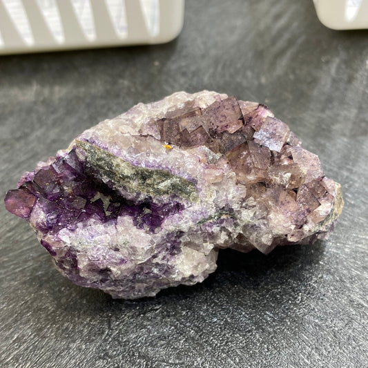 A rough, unpolished fluorite crystal rock from The Crystalary's LIVE-autisticmedium-5/11/24 collection sits on a dark textured surface. The rock displays various shades of purple, white, and gray, with a noticeable band of purple running through the center. Its uneven surface features jagged edges and small cubic formations that are reminiscent of minerals commonly found in Canada.