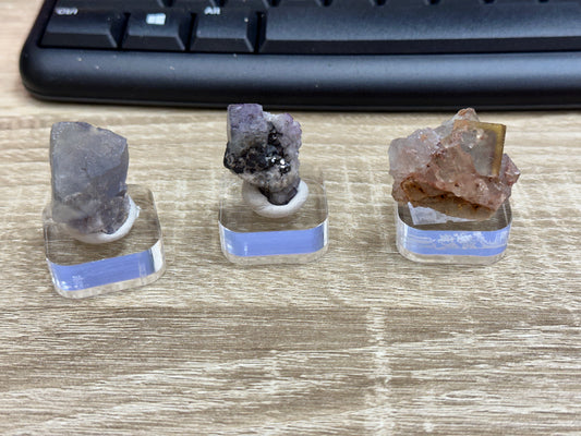 Three mineral samples from The Crystalary's LIVE- Bast- 5/11/24 collection are displayed on clear stands, arranged on a wooden surface. Among them is a striking fluorite from El Hammam. Positioned in front of a black computer keyboard, each mineral specimen flaunts its unique color and texture, showcasing an intricate crystalline structure worthy of Lady Annabella’s collection.