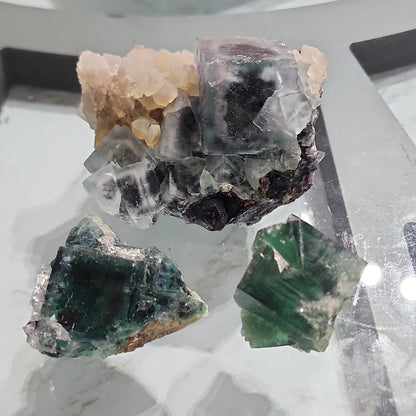 A close-up photo of three fluorite crystals from the Okorusu Mine, featured in The Crystalary's LIVE- Bast1776- 4/15/24 collection, placed on a reflective surface. The crystals exhibit a stunning range of colors, including green, purple, and clear sections, and have distinct geometric shapes that showcase their natural beauty.