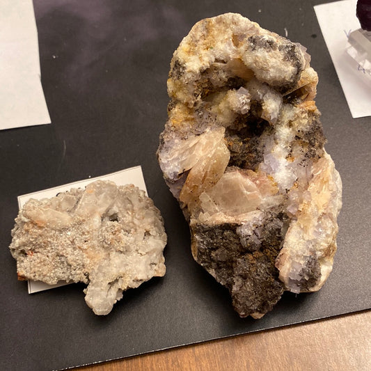 Two mineral specimens displayed on a dark surface. The larger specimen, from the LIVE- Casper- 11/21/23 collection by The Crystalary, is rich with Goethite and has a rough, crystalline texture with white, gray, and brown hues. The smaller specimen to its left is more compact and predominantly Calcite white with some translucent areas.