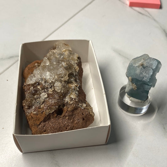 Displayed in a white rectangular box on the left is a brown rock with clear crystalline formations. To the right, there stands a smaller blue-green Fluorite crystal placed on a small, transparent pedestal. Both items from The Crystalary are showcased on a light-colored surface.