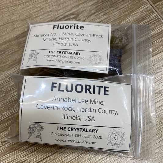 Two small plastic bags lie on a wooden surface, each containing pieces of fluorite and labeled with details. The labels indicate the source mines and locations: Minerva No. 1 Mine and Annabelle Lee Mine, Cave-In-Rock, Hardin County, Illinois, USA. These items are part of The Crystalary's "LIVE- CheerBow- 6/6/24" collection.